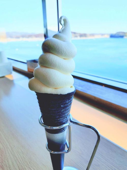 Soft-serve vanilla ice cream cone in a dark wafer cone, held in a metal stand on a wooden table by a window overlooking water.