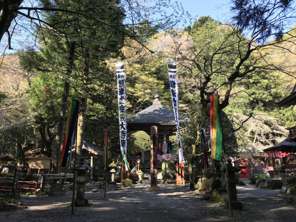 Shinto shrine courtyard with tall vertical banners, stone lanterns, and a small wooden building surrounded by trees on a sunny day.