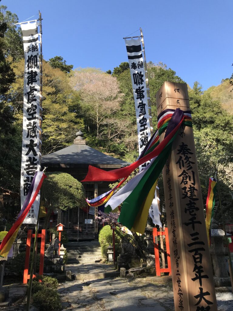 Shinto shrine entrance with tall vertical banners and colorful fabric streamers, stone pathway, and a wooden gate in a forested setting.