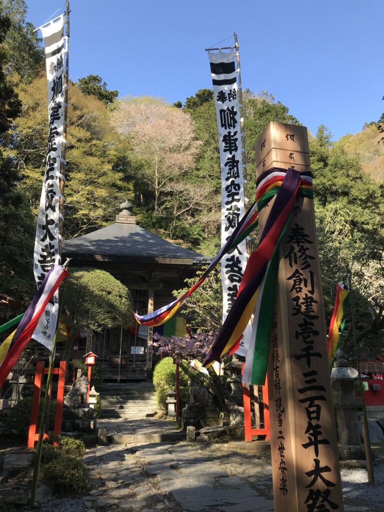 Temple courtyard with stone path, banners, and a wooden shrine surrounded by trees on a sunny day in Japan.