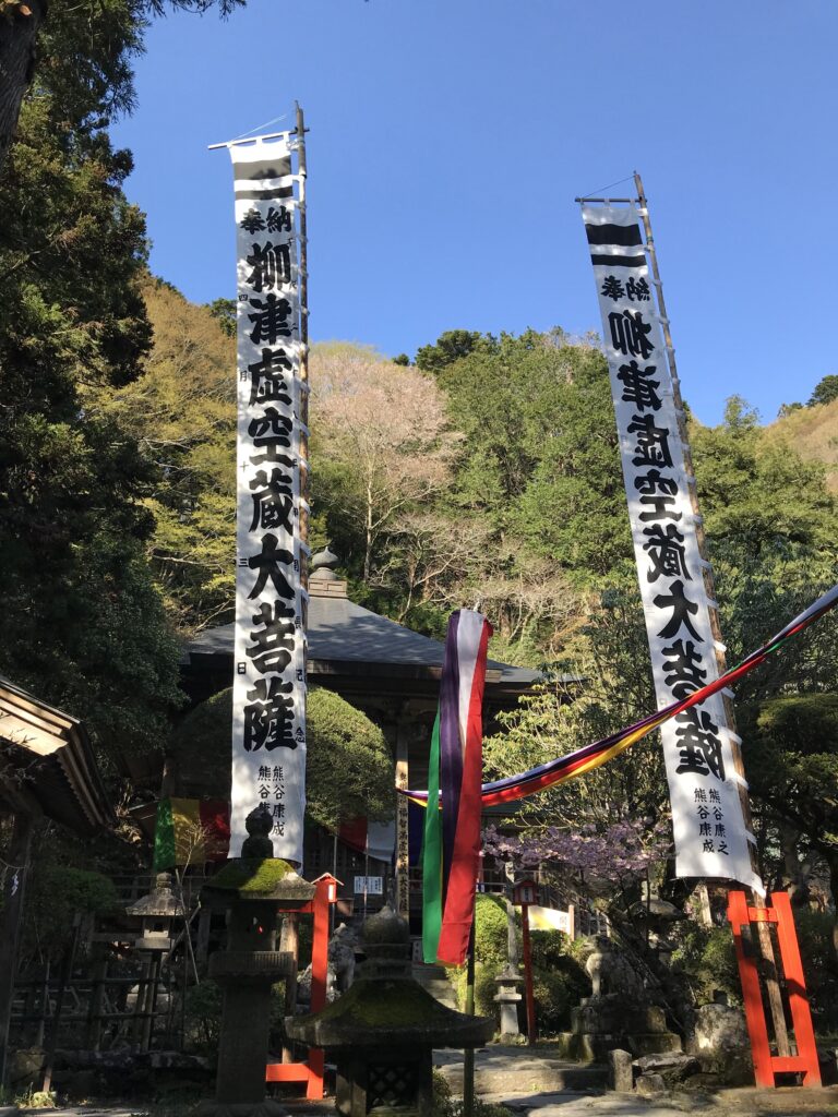 Shinto shrine courtyard with tall white banners bearing black kanji, stone lanterns, and colorful ribbons against a blue sky.