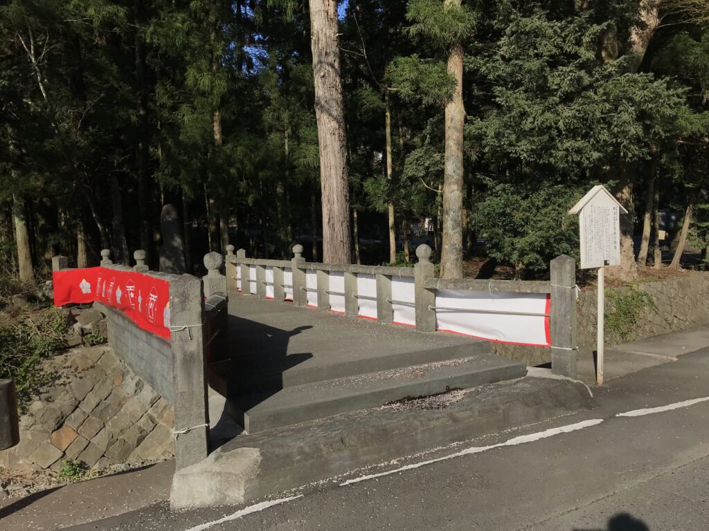 Stone pedestrian bridge with red fabric banners along the railing, leading into a forested area with tall trees and a white signboard nearby.