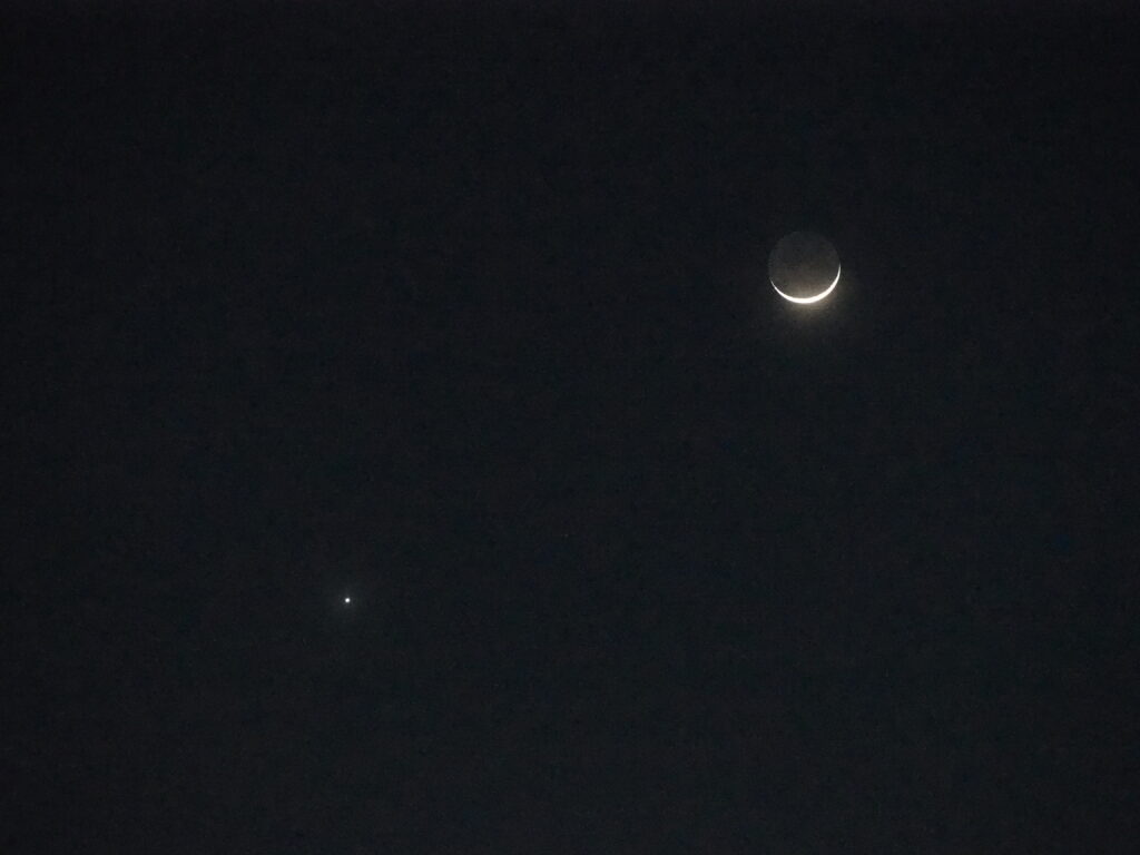 Crescent moon visible in a dark night sky with a bright planet to the lower-left.