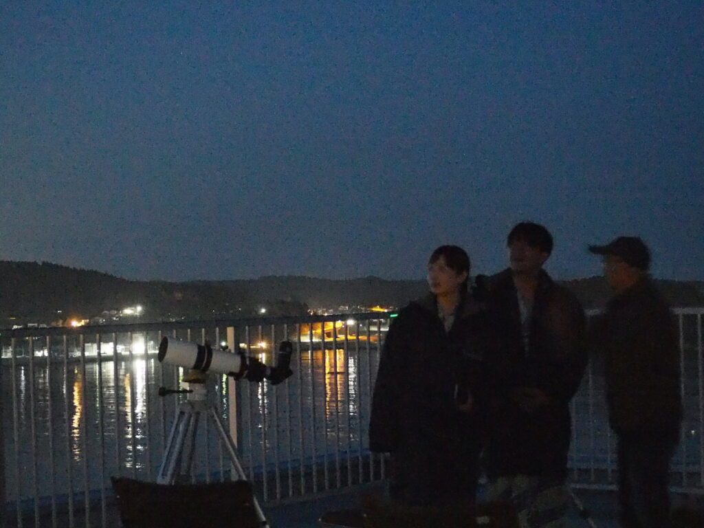 Three people stand by a railing at night beside a telescope on a tripod, overlooking a river with city lights.