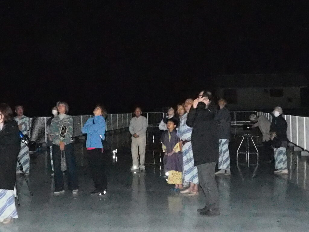 Group of people standing on a rooftop at night, looking upward as if stargazing; railing and equipment visible in the background.