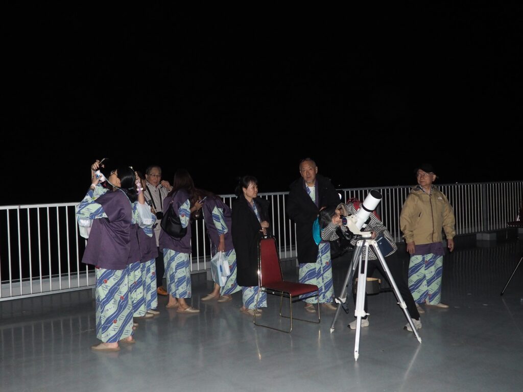 Group of people in matching blue patterned robes on a rooftop at night, some taking photos while a telescope observes the sky.
