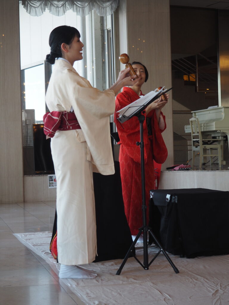 Two women in traditional kimonos perform on a stage; one holds a wooden percussion instrument near a music stand with sheets of music nearby.