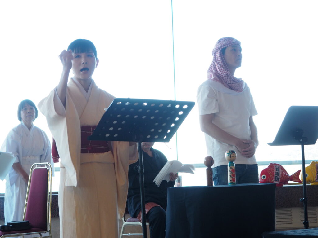 People in traditional robes performing on a stage with music stands; woman raises a hand as she sings, man wearing a keffiyeh looks on; colorful props on the table nearby.