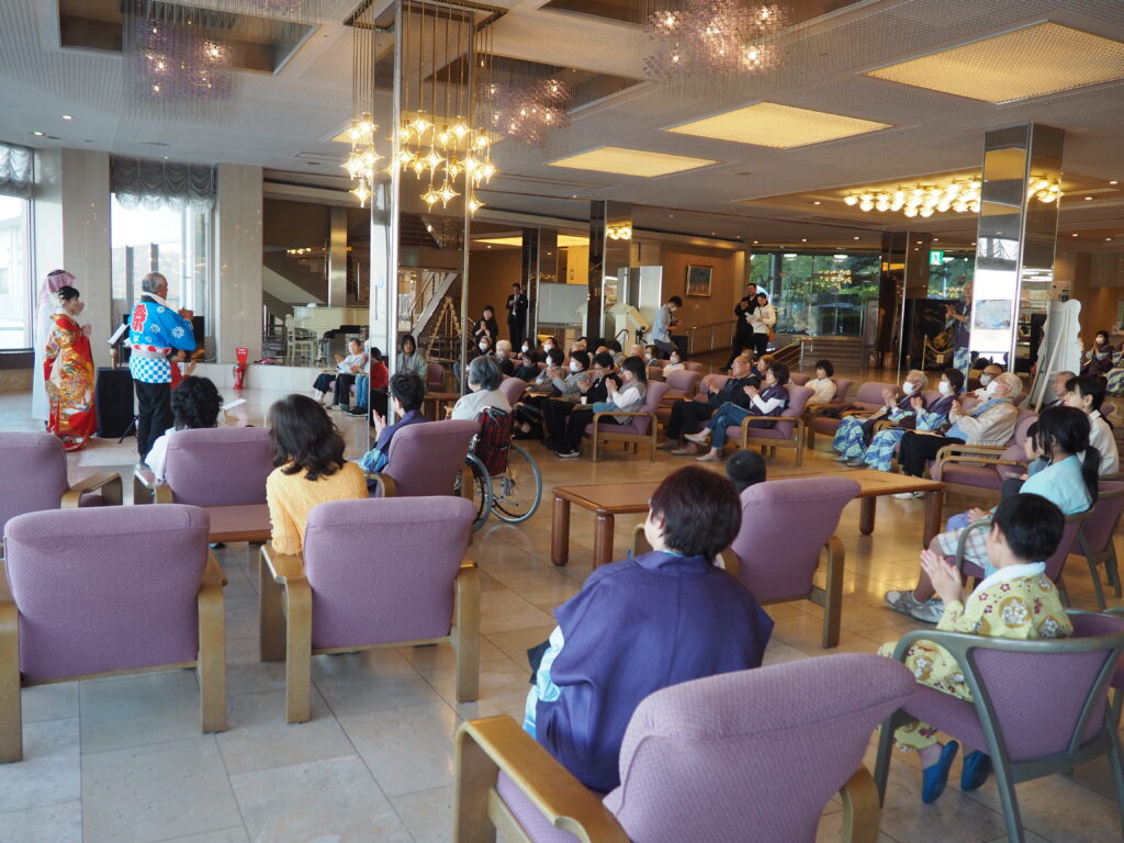 Live cultural performance in a hotel lobby with performers in colorful kimono facing an audience of seated attendees.