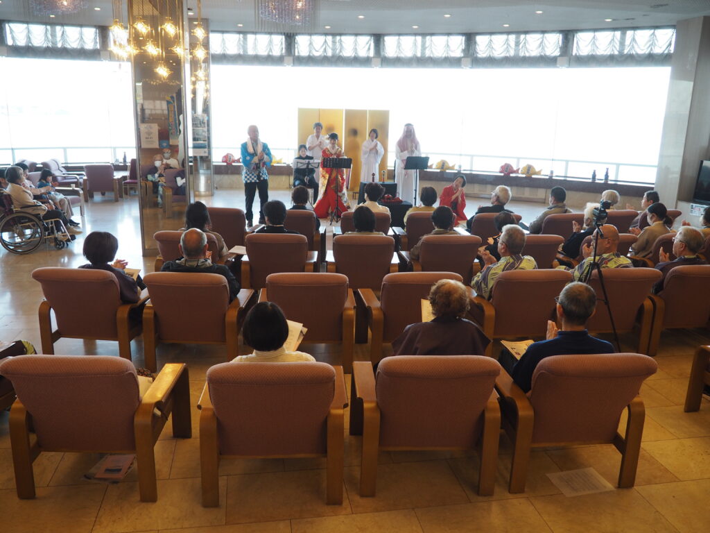 Elderly audience seated in a lounge watching a live performance by performers in traditional Japanese attire on a small stage with a gold screen.