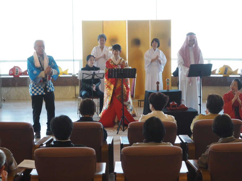 Group of performers in traditional Japanese attire on stage with a gold folding screen, audience seated in front.