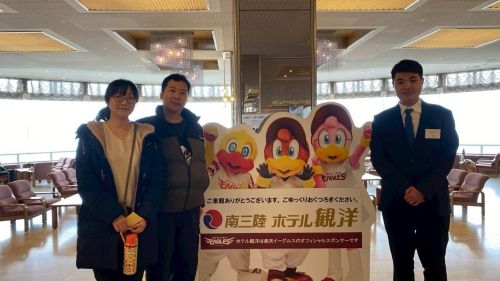Four people pose in a hotel lounge beside a cartoon mascot cutout and a sign with Japanese text reading Minamisanriku Hotel Kanyo, winter clothing on display.