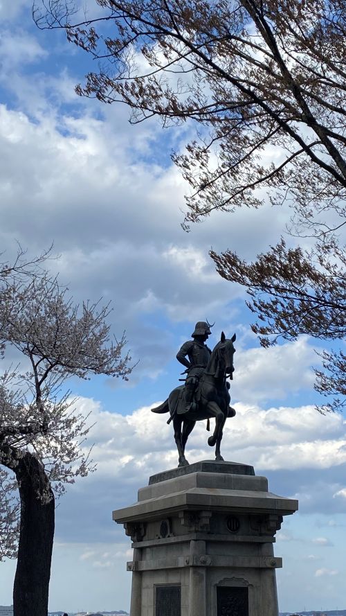 Bronze equestrian statue atop a stone pedestal, seen from below, with bare trees and a blue, partly cloudy sky.