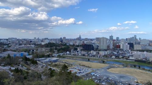 A distant city skyline under a blue sky with scattered clouds, viewed from a hill above a parking lot and river.