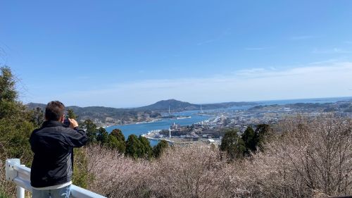 Person in a dark jacket at a railing, photographing a harbor town with boats and hills under a blue sky.