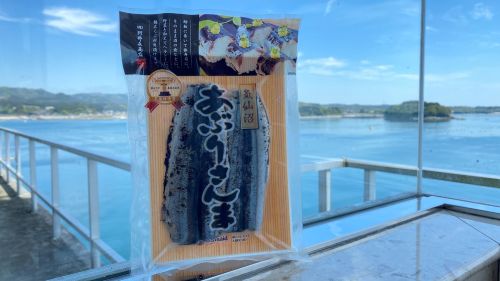 Packaged Japanese snack in a sealed blue-labeled bag on a wooden backing, with the blue sea and railing in the background.
