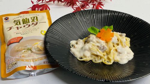 Plate of pasta in creamy white sauce with a decorative orange carrot-shaped garnish, served on a dark plate, beside a yellow package labeled 気仙沼チャウダー (Kesen-numa Chowder).