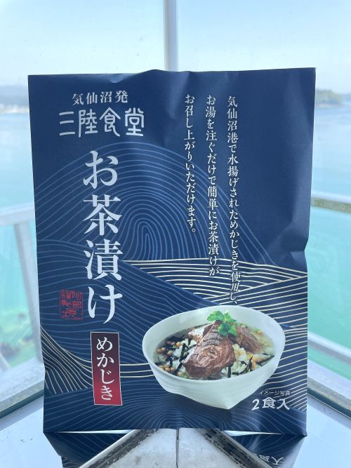 Blue package labeled 三陸食堂 お茶漬け with a bowl of ochazuke (fish over rice) pictured; seascape visible through window behind.