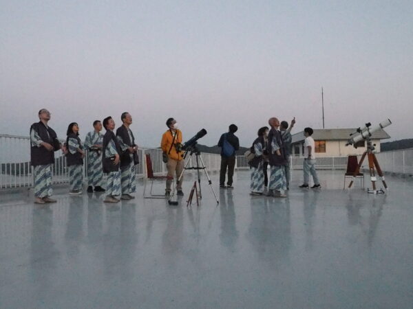 Group of people in yukata on a rooftop, standing with telescopes and equipment, looking up at the sky at dusk/faint light