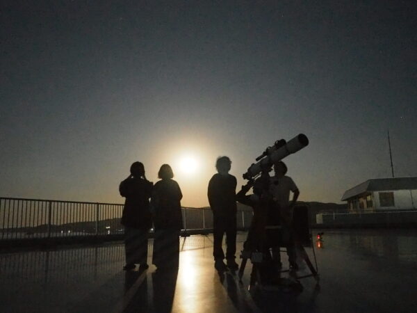Silhouetted group on a rooftop with a telescope, looking up at a starry night sky with a bright horizon glow behind them.
