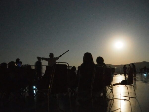 Silhouetted group of people on a rooftop or terrace at night, with a bright moon or light source in the sky behind them.