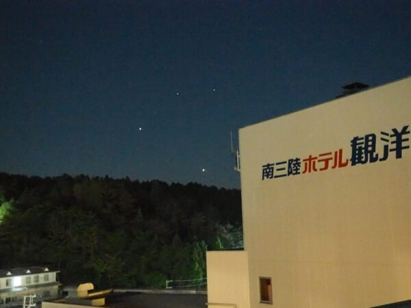Nighttime exterior of a beige building with large Japanese signage on the right, dark sky with a few stars, and trees in the foreground.