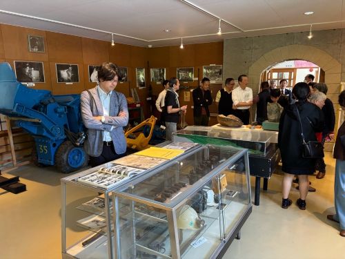 Museum exhibition room with visitors examining glass display cases and a large blue industrial machine on the left.