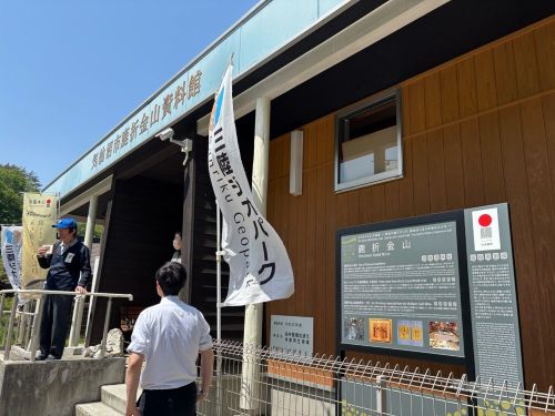 People stand outside a wooden information center with Japanese signage and flag banners on a sunny day.