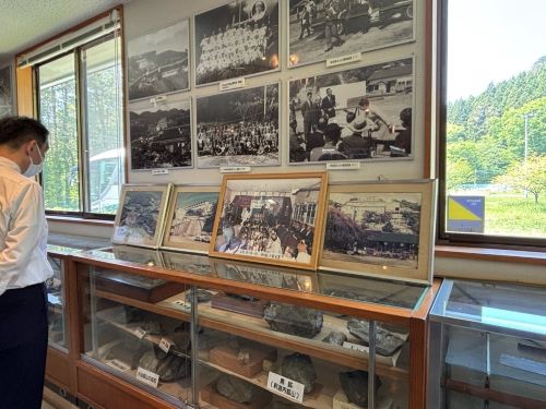 Man in a white shirt and mask viewing a museum display of framed photos and a glass cabinet filled with artifacts.