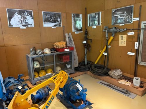 Museum display of vintage mining equipment: blue and yellow machinery, helmets, pipes, and photos on a wood-paneled wall backdrop.
