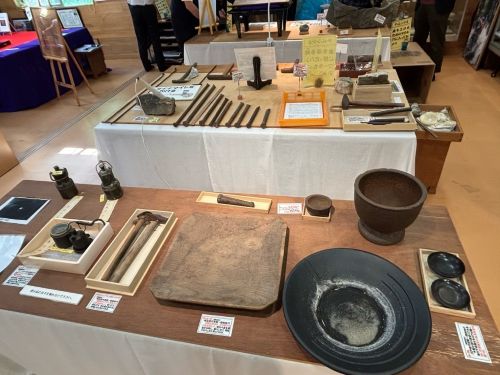 Museum display of antique tools and kitchenware arranged on tables, including bowls, mortars, pestles, and metal implements.