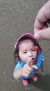 Young child in a pink hat reaching up to hold a small red cherry on a stem outdoors on dirt ground, wearing a blue shirt and red shoes with bear faces.