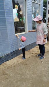 Child in a pink hat reaches up to a window ledge with a stick, guided by a woman in a pink apron outside a blue tiled building.