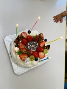 Birthday cake topped with strawberries, blueberries, and mint, with green, pink, and yellow candles lit; a child's hand rests near the edge of the table.