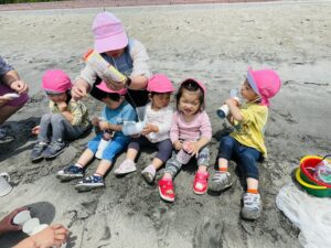 Group of young children wearing pink hats sit on a rocky beach, an adult hands out cups and snacks. -