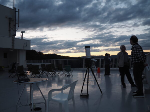 Rooftop deck at dusk with folding chairs, a telescope on a tripod, and a cloudy sky; people on the right wearing masks.