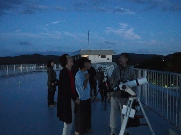Group of people on a rooftop at dusk gathered around a large telescope setup outside a small building with a railing nearby.