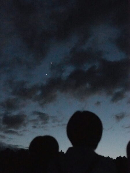Silhouettes of people in the foreground looking up at a dark blue, cloudy night sky with two bright stars/planets.