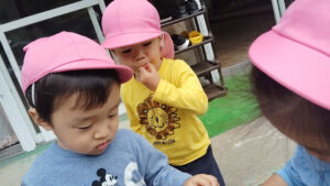 Three kids wearing pink hats stand outdoors, looking down at something, with one in a yellow lion shirt and another in a blue Mickey Mouse shirt.