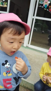 Two preschoolers sit outside near a glass door; one wears a pink cap and is about to eat an orange snack while the other in a yellow shirt watches nearby.