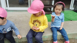 Three preschoolers sit on a concrete step outdoors, wearing pink hats; middle child wears a yellow lion shirt.