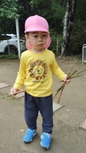 Young child wearing a pink hat and yellow lion-print shirt holds sticks outdoors in a park setting.
