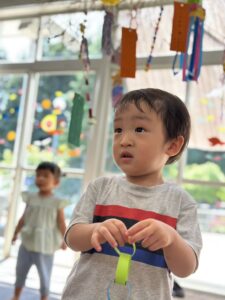 Young child in a gray shirt with red/black/blue stripes holds a bright green ribbon toy in a colorful indoor play area with hanging paper decorations; another child blurred in the background.