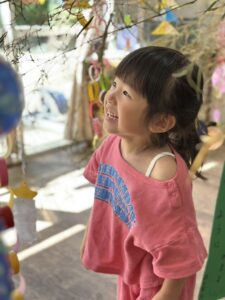 A smiling girl in a pink shirt looks up at colorful hanging decorations outdoors.