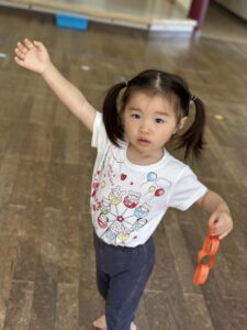 Little girl with pigtails raises her left arm while holding an orange linked-chain on a wooden floor indoors.