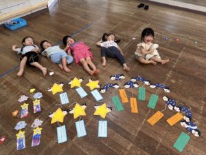 Five children lie on a wooden floor around colorful star-shaped crafts and handwritten tags from a craft activity.",