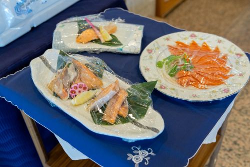 Assorted sashimi platters on a blue tablecloth, wrapped in plastic, with salmon slices and decorative garnishes on plates.