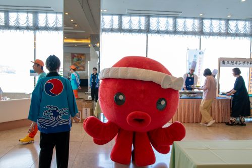 Red plush octopus mascot wearing a white headband stands indoors at an event, with attendees and booths in the background and people in blue happi coats nearby.