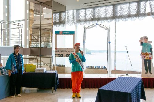 Person wearing a sash and orange pants speaks into a microphone on a stage near a windowed backdrop, with another person and display tables nearby at an event.