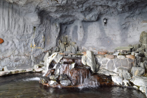 Rocky grotto with grey walls, water cascading over layered stones at the base, and a stone seating wall along the edge.